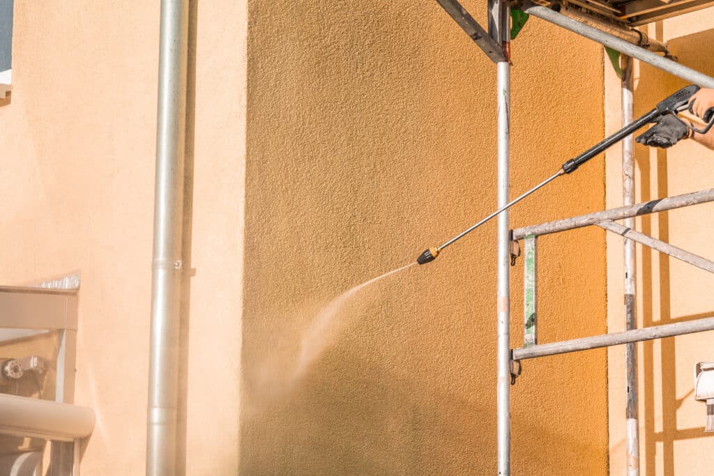 Nettoyage de façade au nettoyeur haute pression Main gantée utilisant un nettoyeur haute pression pour nettoyer une façade texturée orange d'un bâtiment, révélant une zone plus claire. Un échafaudage est visible sur la droite.